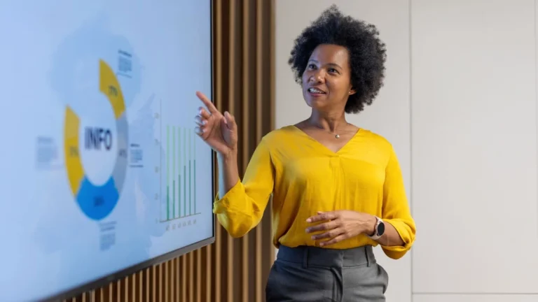 Mulher preta em seus 50 anos vestindo uma blusa amarela aponta para um slide de uma apresentação em inglês